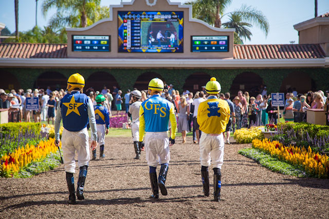 Three jockeys in colorful uniforms walking towards a racetrack with spectators in the background.