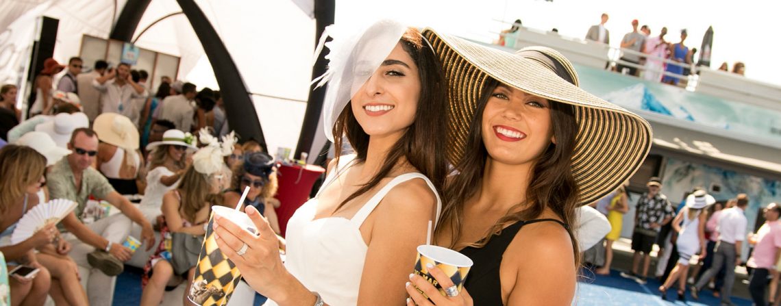Two women wearing large hats and holding drinks at a social event with attendees in the background.