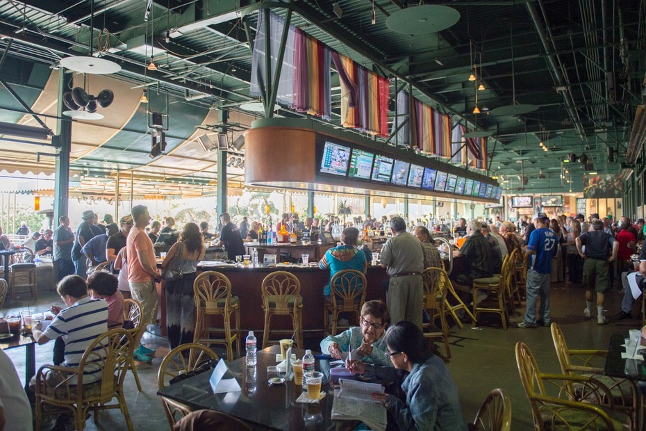 A bustling restaurant interior featuring a bar area, patrons enjoying drinks, and multiple screens displaying events.