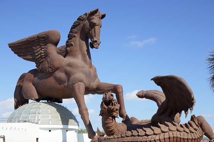 Large sculpture of a winged horse confronting a dragon, set against a blue sky.