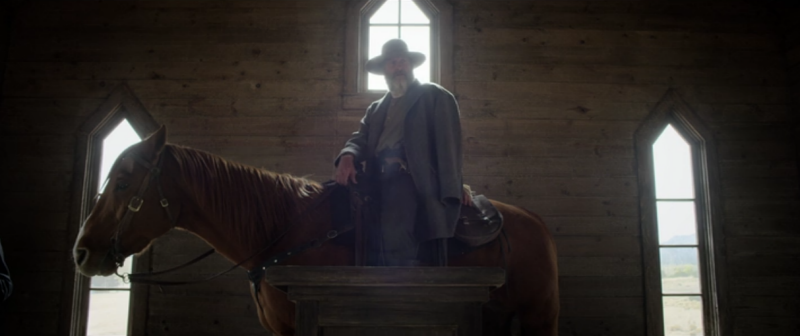 A cowboy in a long coat standing on a wooden platform beside a horse inside a rustic barn with arched windows.
