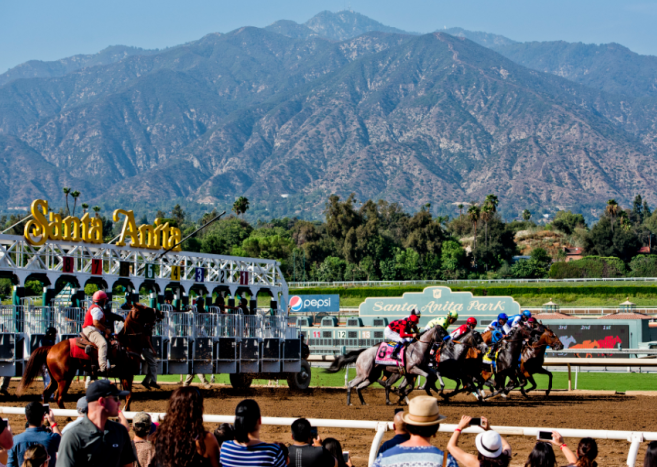 Horses and jockeys racing at Santa Anita Park with mountains in the background.