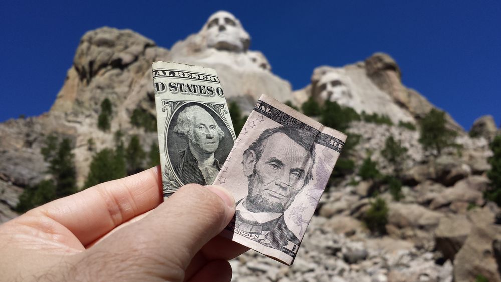 A hand holding a one-dollar bill featuring George Washington and a five-dollar bill featuring Abraham Lincoln in front of Mount Rushmore.