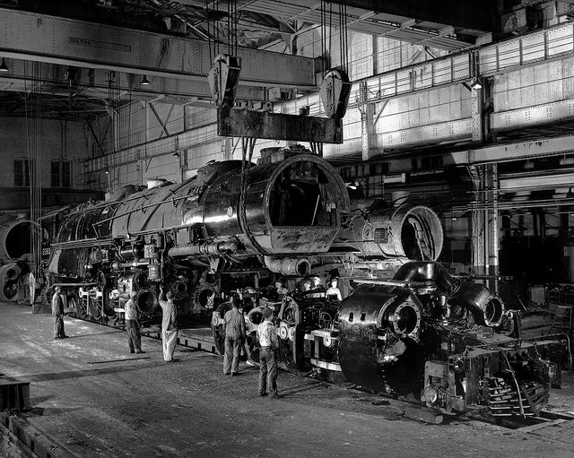 A steam locomotive being assembled in a factory with workers present around it.