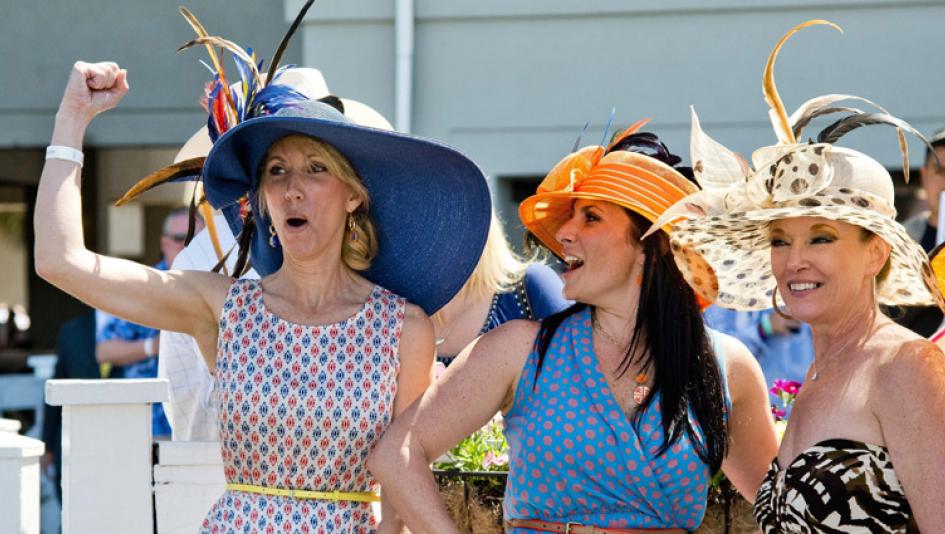Three women wearing colorful and decorative hats, expressing excitement at an outdoor event.