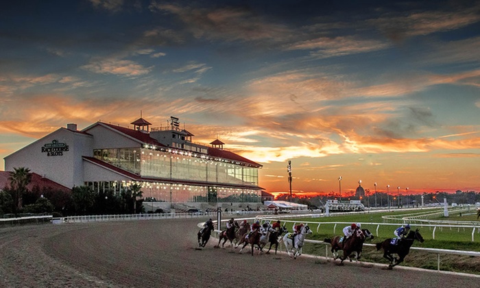 Horses racing on a track with a grandstand and sunset in the background.