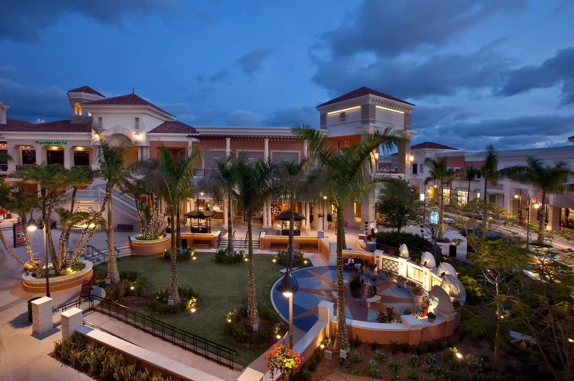 An outdoor shopping center featuring palm trees and architectural structures under a dusk sky.