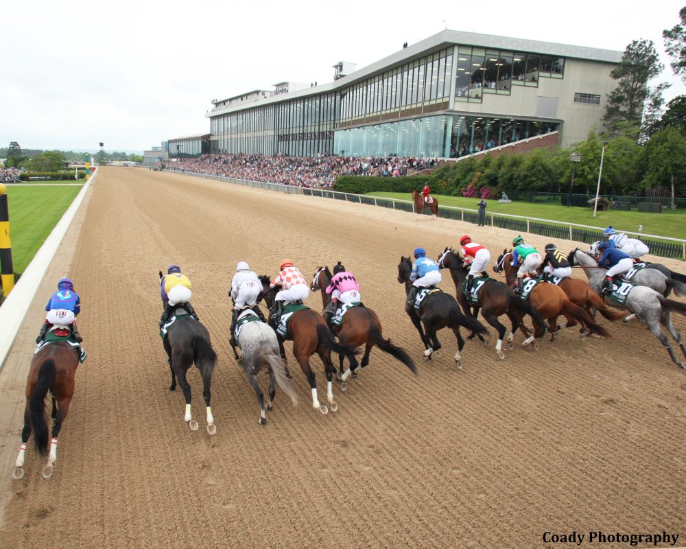 Horses racing on a dirt track with jockeys in colorful silks at a racecourse.