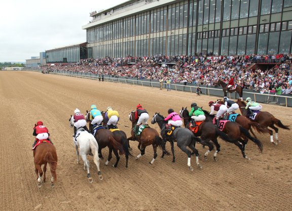 A group of horses and jockeys racing on a dirt track with spectators in the background.