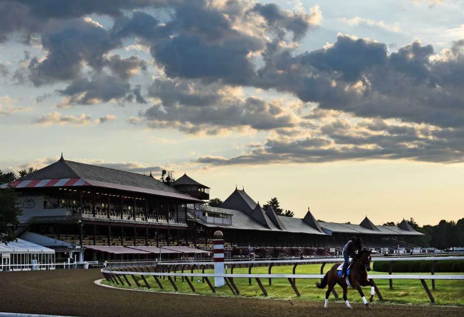 A horse walking along the track at Saratoga Race Course with grandstands in the background under a cloudy sky.