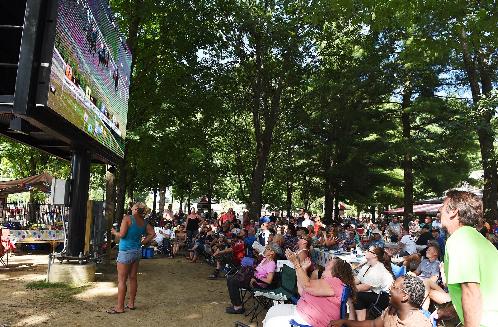 Crowd watching a large screen at an outdoor event surrounded by trees, showcasing a lively atmosphere with engaged spectators.