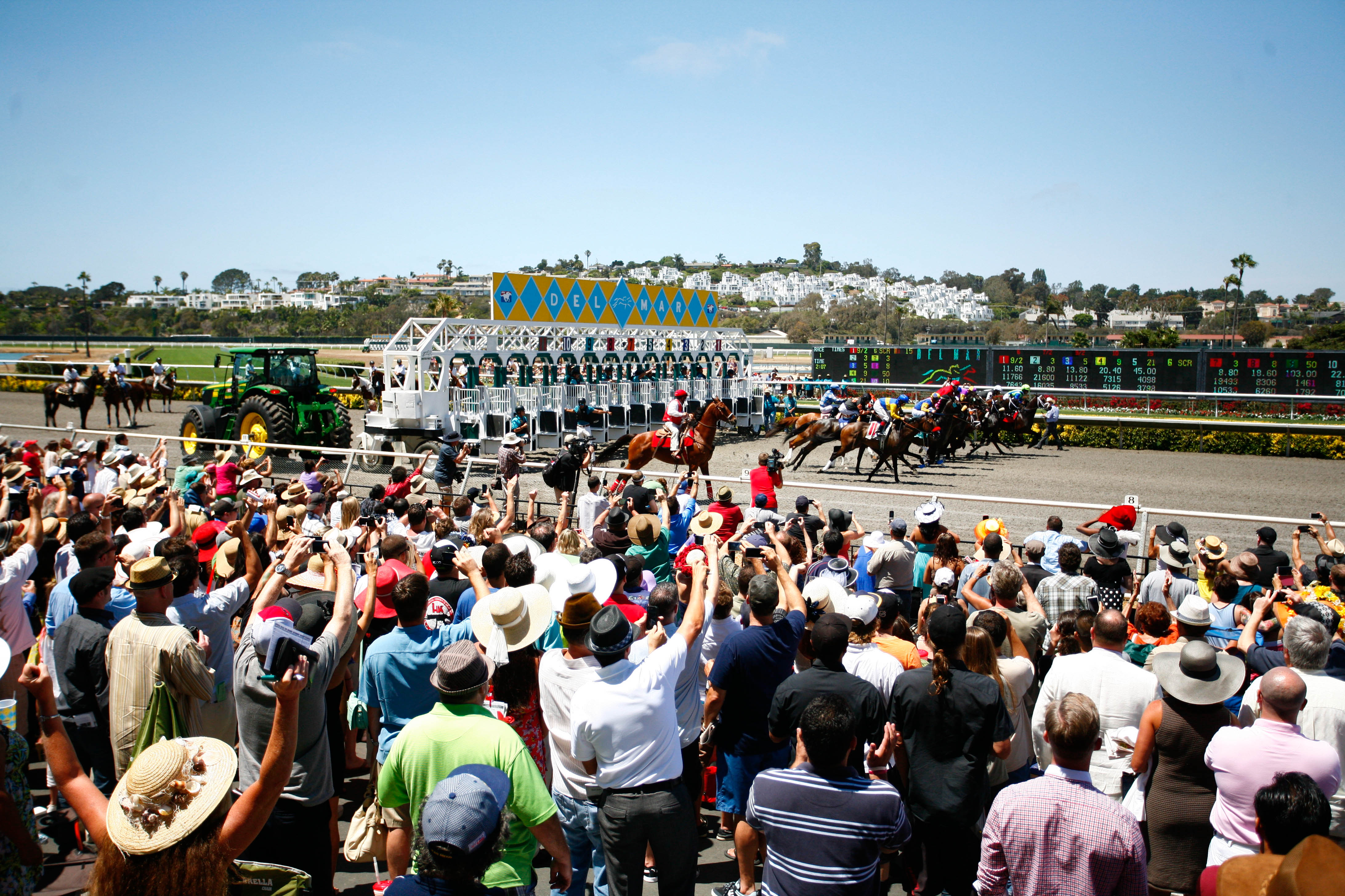 A group of horses exiting the starting gate at a horse racing event with spectators in the foreground.