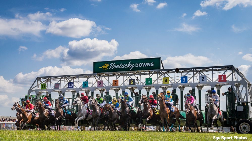 Horses and jockeys racing from the starting gate at Kentucky Downs under a blue sky.
