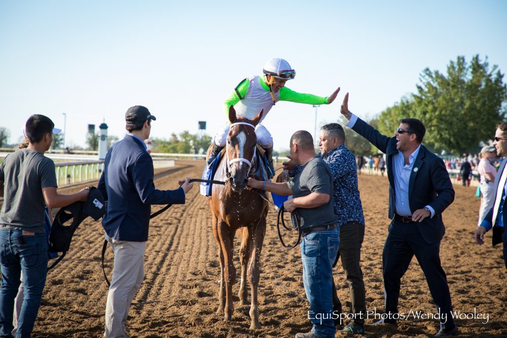 Jockey in bright attire celebrating on a horse with a group of people at a race track.