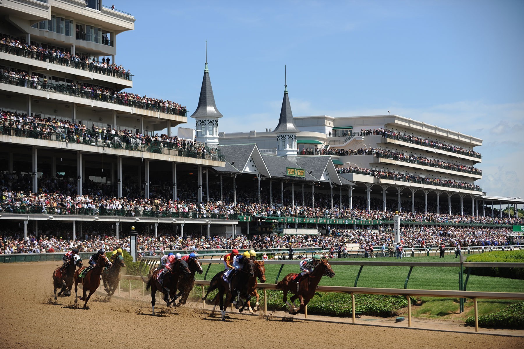 Horses racing at Churchill Downs with spectators in the grandstands during a sunny day.