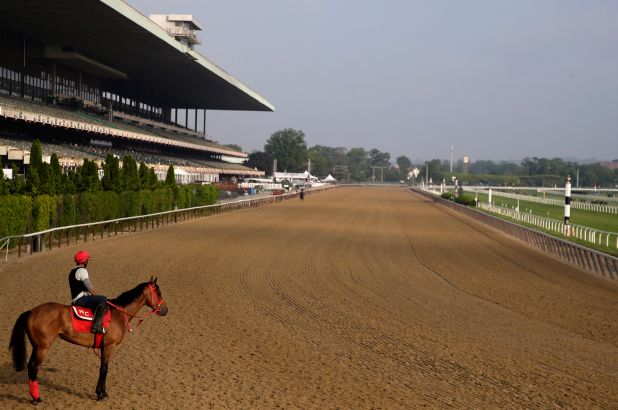 A horse with a jockey in red gear standing on the dirt racetrack at Belmont Park.