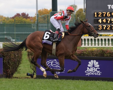 A jockey in red and white attire riding a thoroughbred horse during a race at the Breeders' Cup event.
