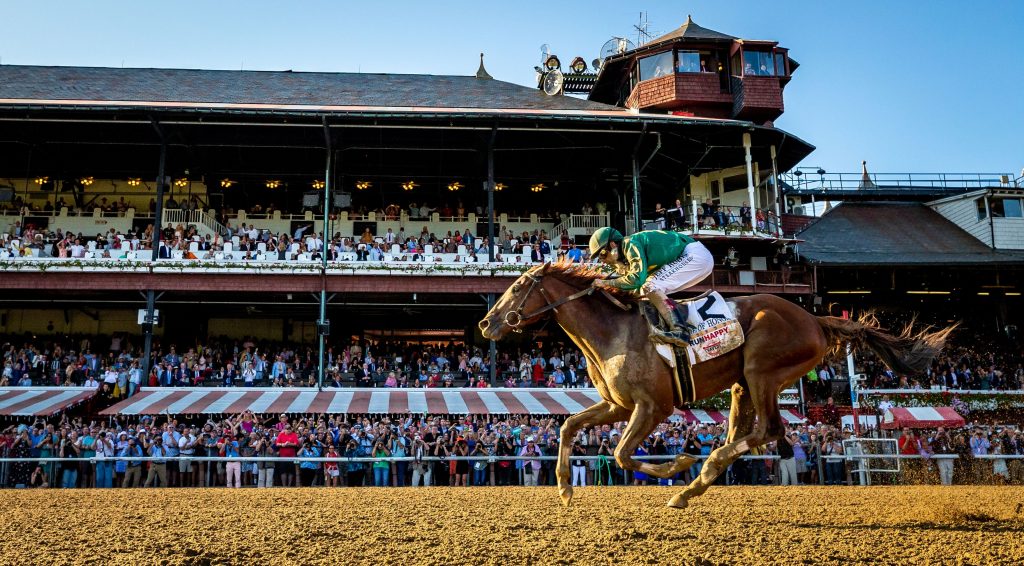 A racehorse with a jockey in green attire running on a dirt track at Saratoga Race Course.