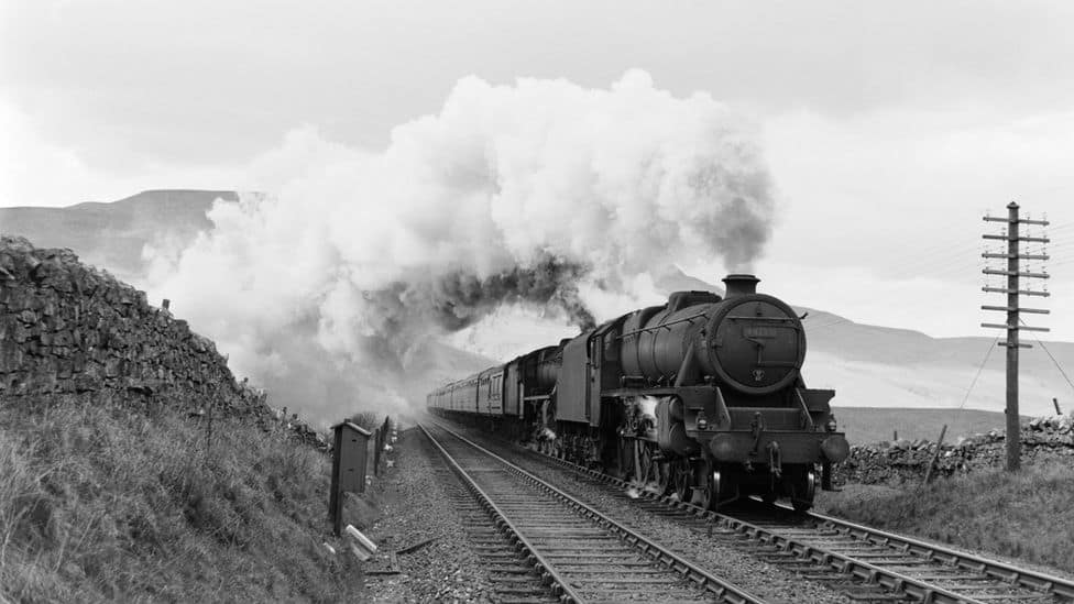 A vintage steam train releasing steam while traveling on railway tracks, surrounded by scenic hills and a cloudy sky.