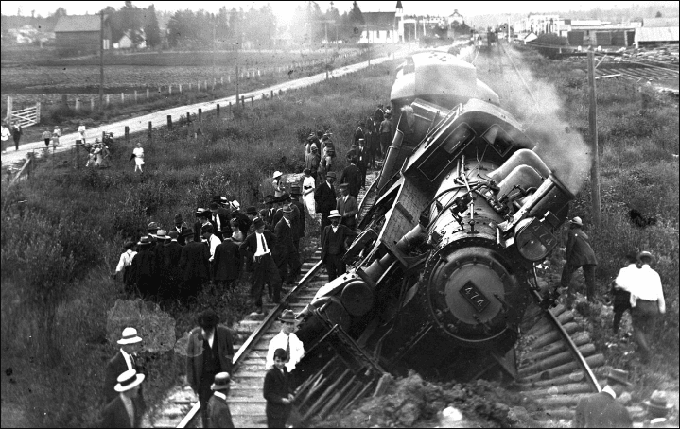 Black and white image of a historic train derailment with people gathered around the overturned locomotive.